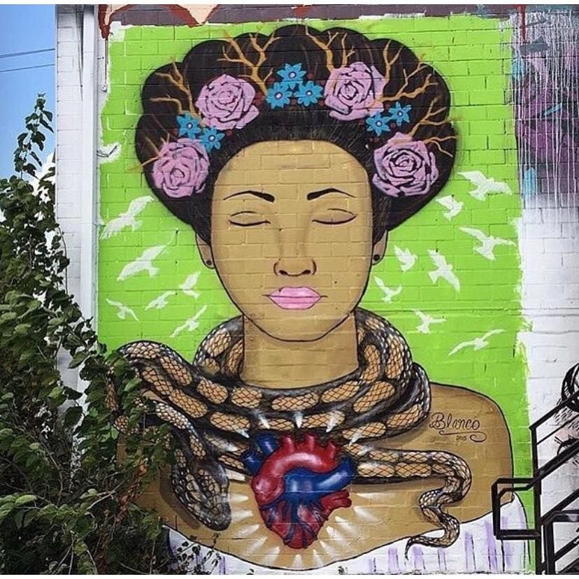 Mural of a woman with closed eyes crowned with roses, wrapped by a snake, with an anatomical heart at her chest, on a lime-green brick wall