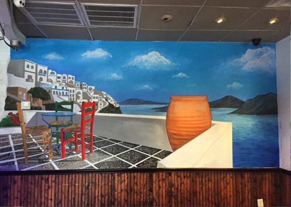 Restaurant wall painted as a Greek island terrace with whitewashed houses, red chairs, a clay urn and the sea